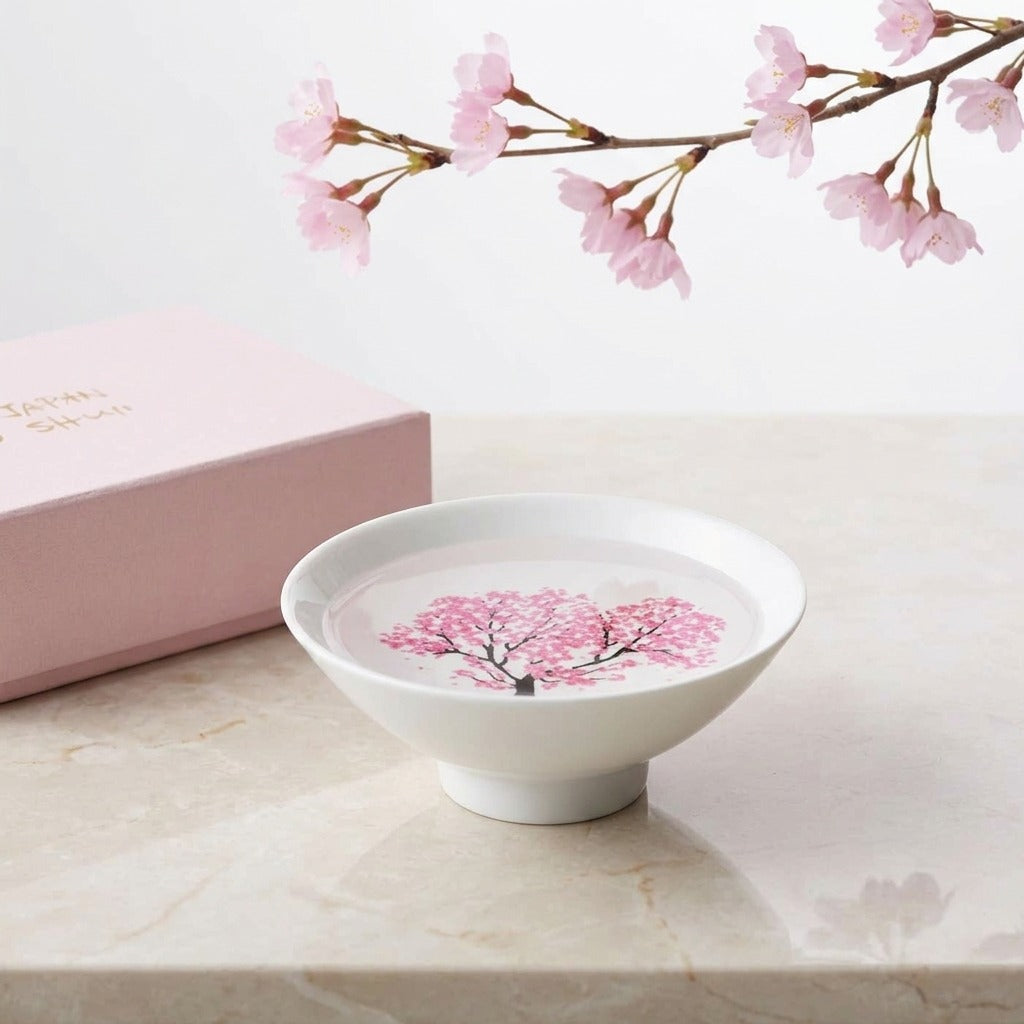 EXTORIL HIYAZAKURA ONE: a single white Mino ware sake cup on a marble table, cherry blossoms bloomed pink by a cold pour, styled with a blossom branch and a box. Only the cup is included.