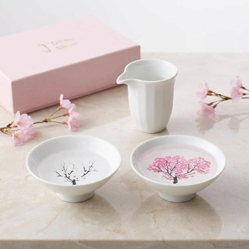 EXTORIL HIYAZAKURA PAIR: two white Mino ware sake cups on a marble table, one plain at room temperature and one bloomed pink after a cold pour, styled with a white sake pitcher, a box and blossom. Only the two cups are included; the pitcher is not.