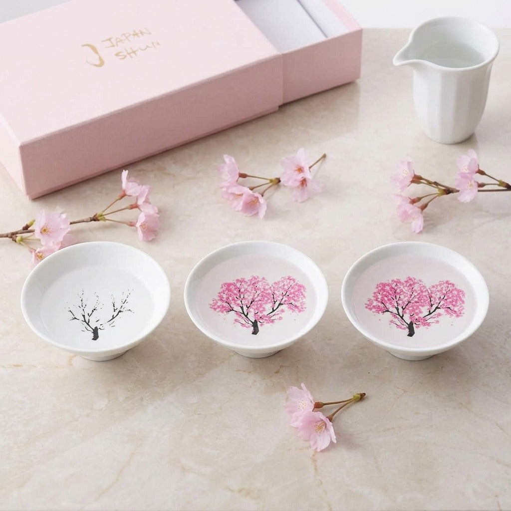 EXTORIL HIYAZAKURA TRIO: three white Mino ware sake cups in a row on a marble table, one plain and two bloomed pink after a cold pour, styled with a white sake pitcher, a box and blossom. Only the three cups are included; the pitcher is not.